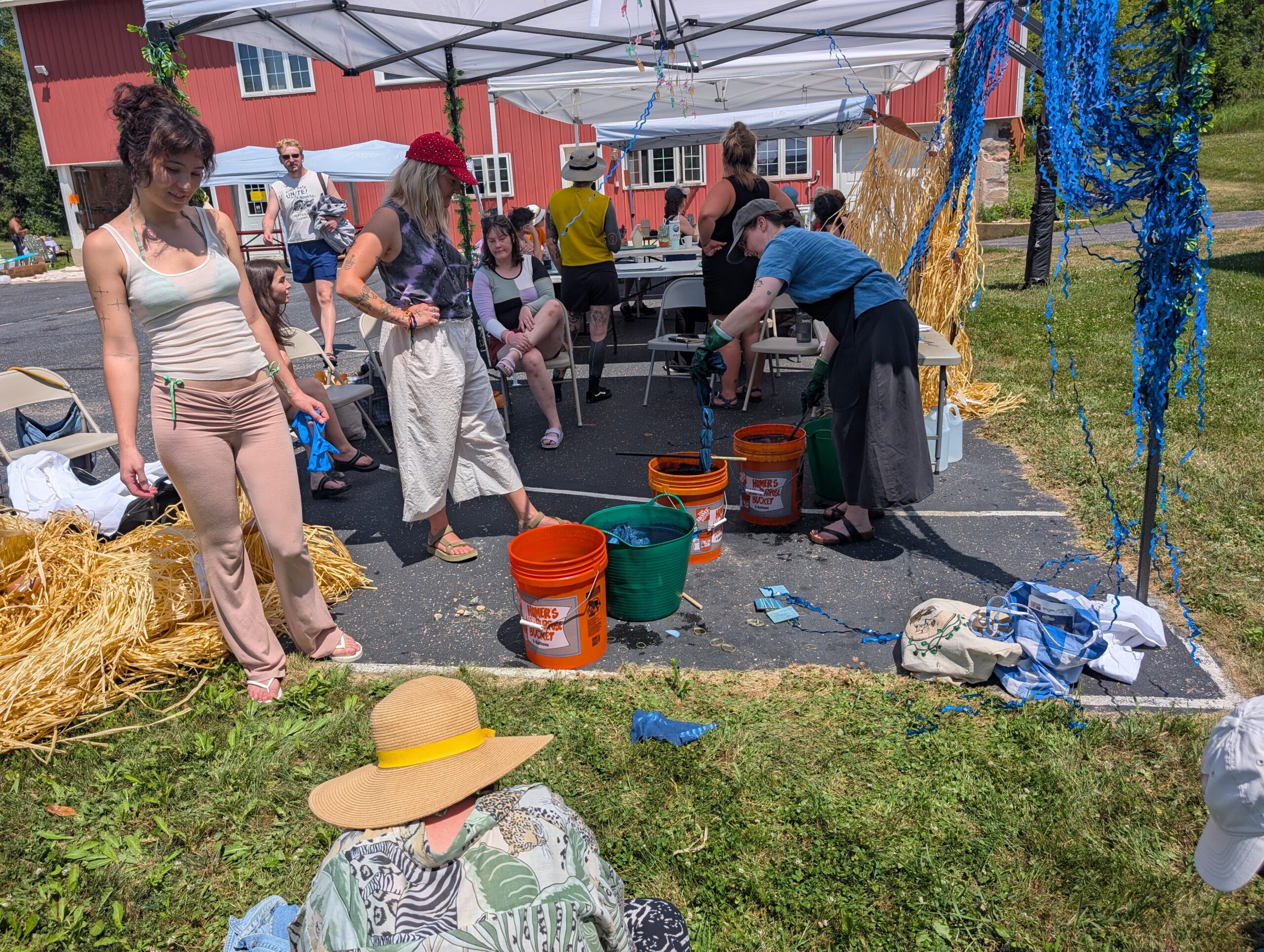 Participants gather under outdoor canopy tents for an indigo dyeing workshop, with dyed fibers hanging to dry and buckets of dyeing materials arranged on the ground.