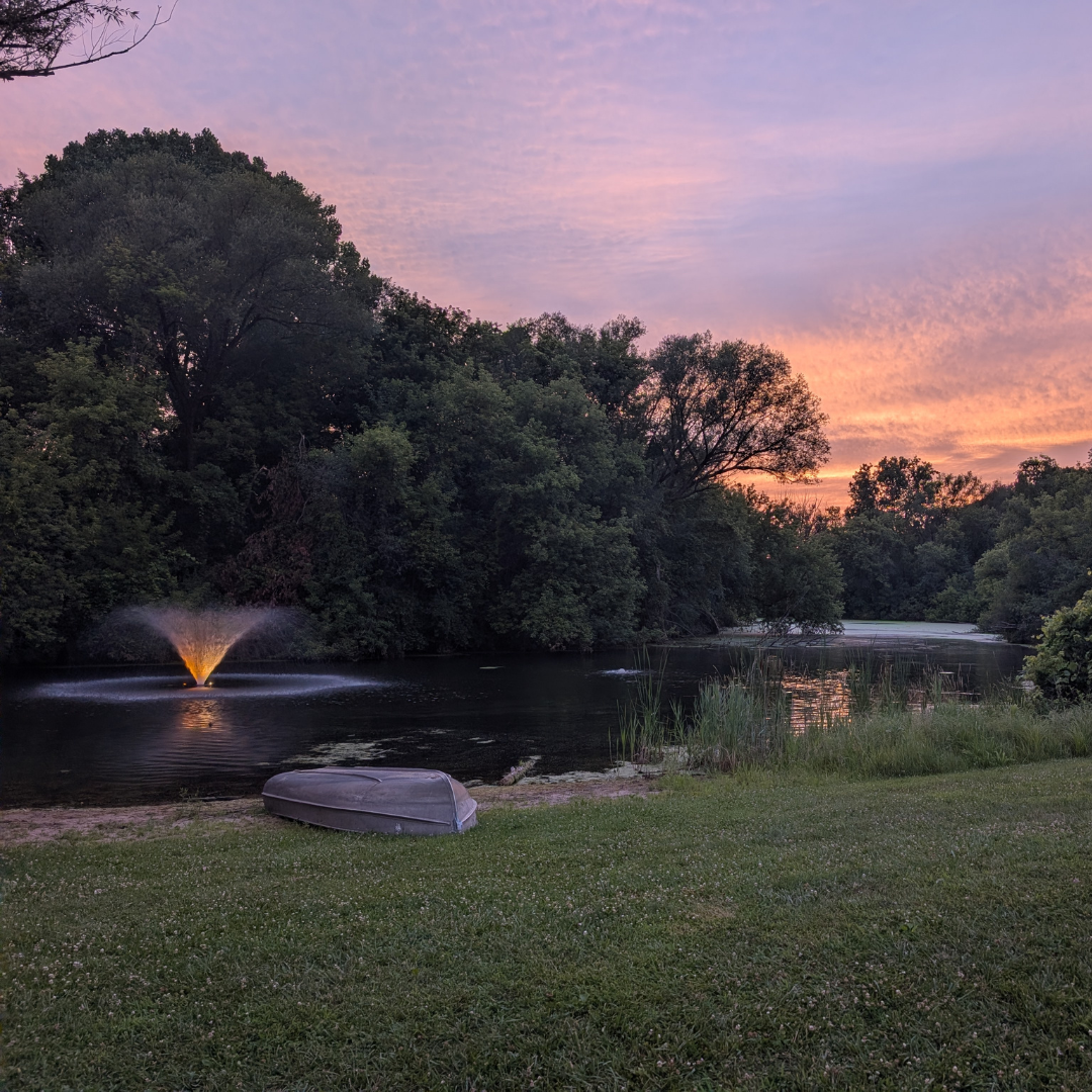 Pond surrounded by trees at sunset, with a small illuminated fountain and a rowboat resting on the grassy shore.