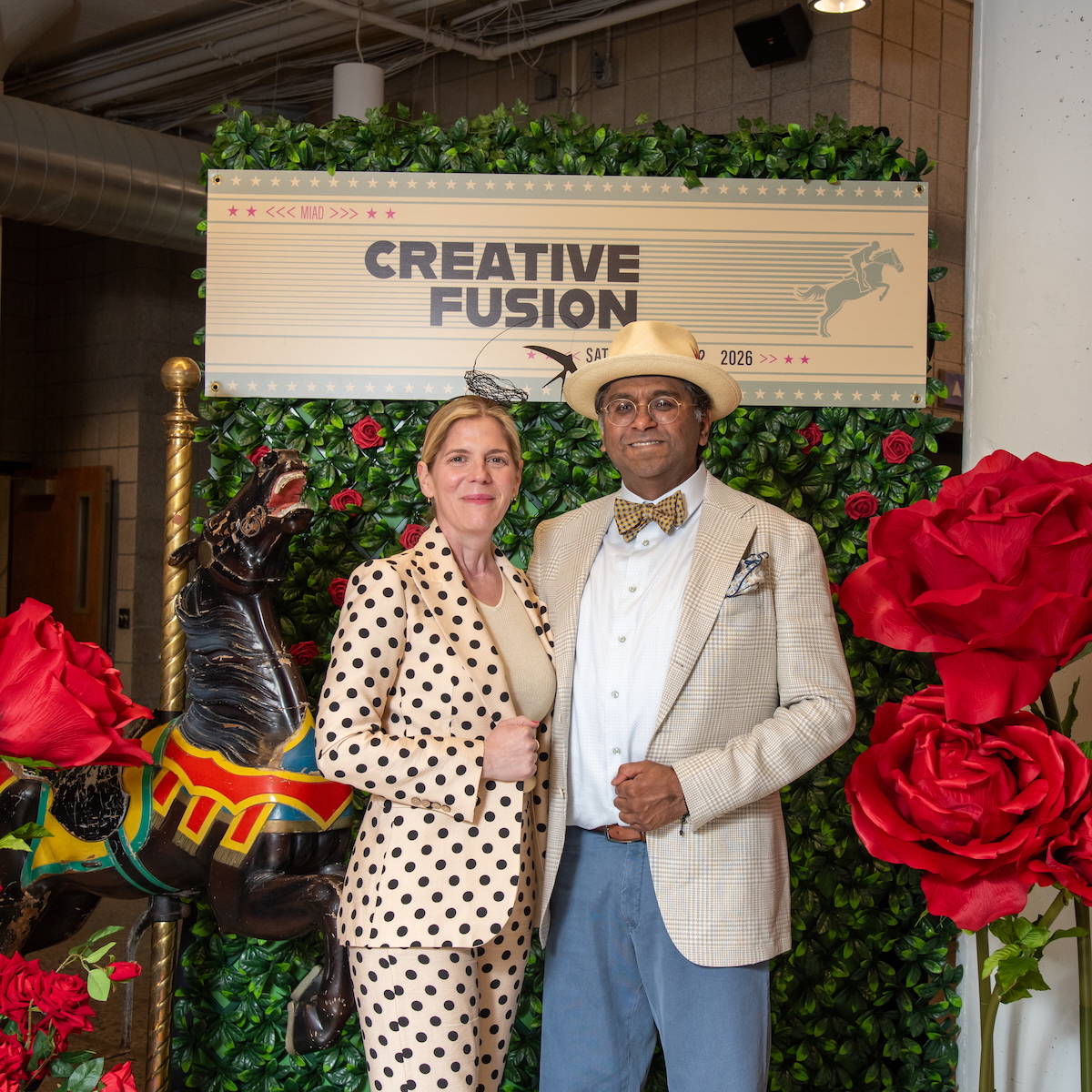 Two event attendees pose in front of a Creative Fusion backdrop decorated with greenery and oversized red roses. A carousel horse figure stands beside them, and a sign above reads “Creative Fusion.”