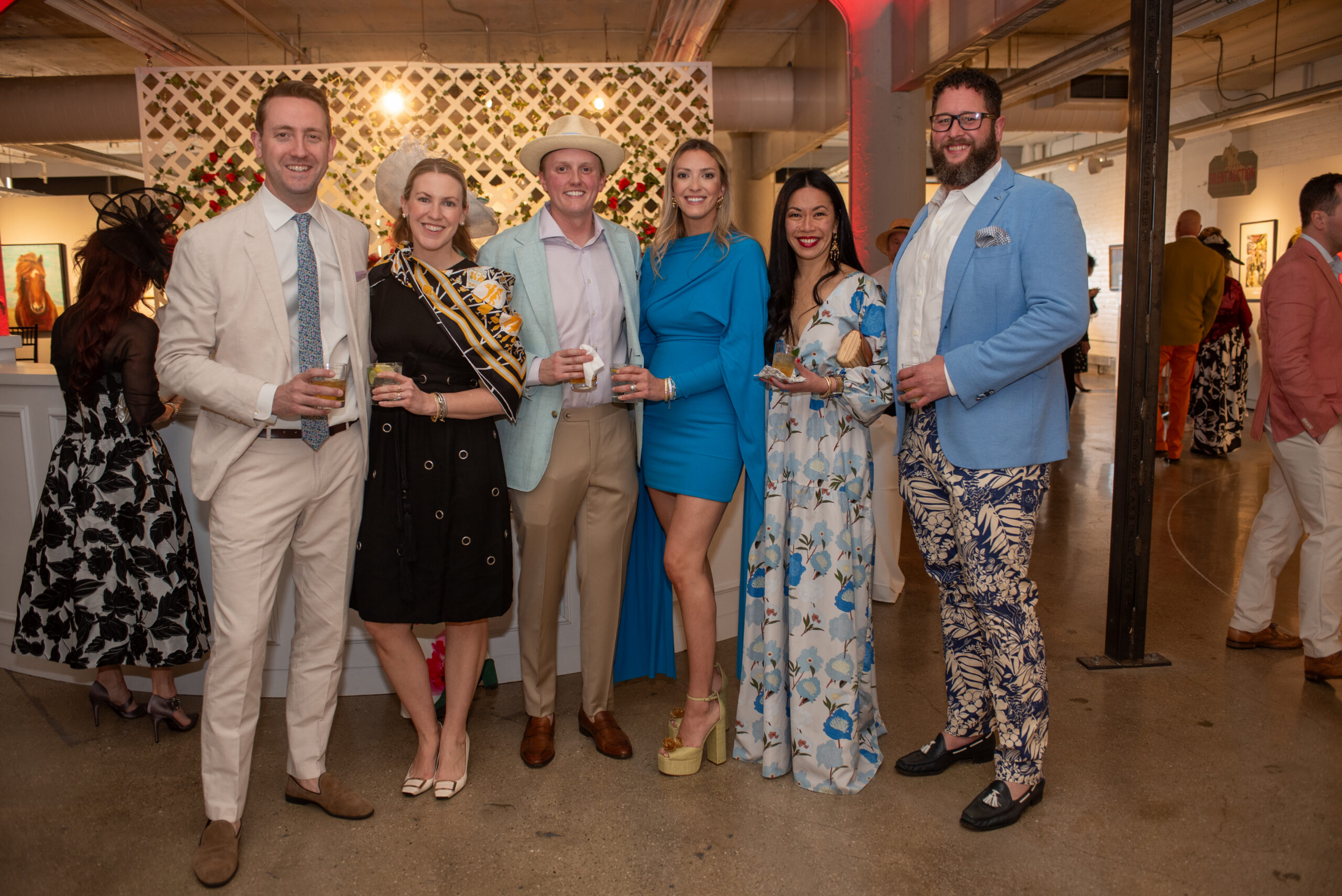 Six attendees pose together at MIAD's Creative Fusion gala inside an event venue. The group stands in front of a decorative lattice backdrop with flowers while holding drinks and wearing colorful formal attire.