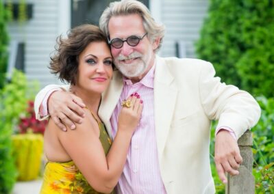 Two people pose together in a garden setting in front of a house. One leans against a wooden fence while both face the camera amid colorful flowers and greenery.