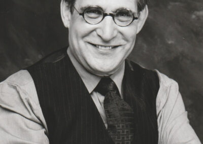 Black-and-white portrait of a person wearing round glasses, a dress shirt, tie, and pinstriped vest, seated with arms crossed.