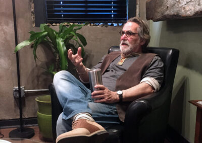 A person wearing glasses sits in a black armchair in a studio or lounge space, holding a metal tumbler and gesturing while speaking. A potted plant, window blinds, and rustic interior details surround the seating area.