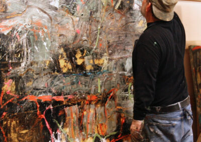 An artist applies paint to a large abstract canvas in a studio. The artwork features layered brushstrokes and splashes of gray, black, white, orange, and gold paint.