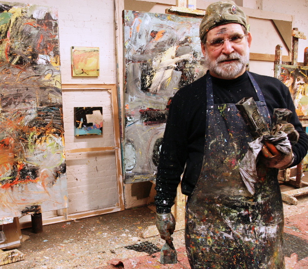 An artist stands in a paint-splattered studio wearing a paint-covered apron and holding painting tools. Large abstract canvases and framed artworks surround the workspace.