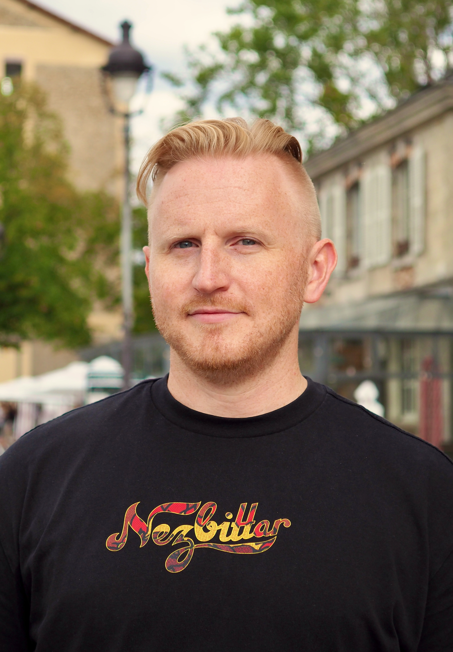 Portrait of a person wearing a black T-shirt, standing outdoors on a city street. Trees, buildings, and a streetlamp appear softly out of focus in the background.