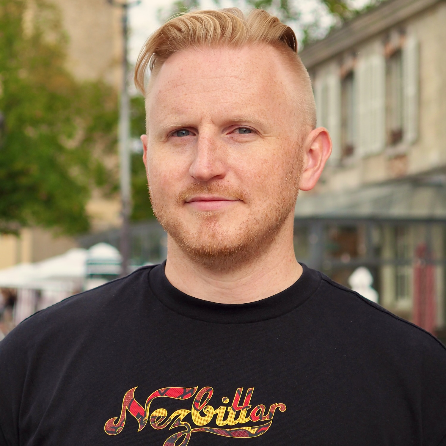 Portrait of a person wearing a black T-shirt, standing outdoors on a city street. Trees, buildings, and a streetlamp appear softly out of focus in the background.