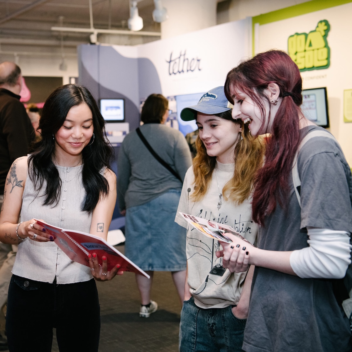 Three prospective students review printed materials while speaking with a representative at a college or education fair. Informational displays and additional attendees are visible in the exhibition space behind them.