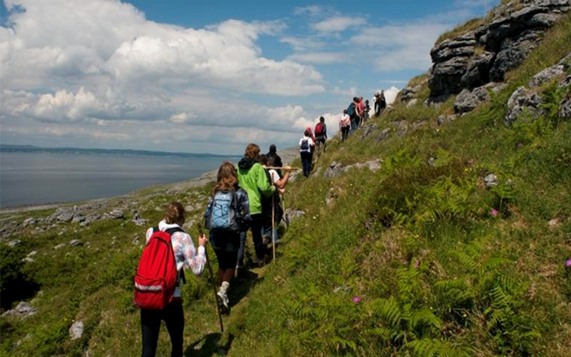 A group of hikers walks along a narrow trail on a grassy hillside overlooking a large body of water. Rocky outcrops and vegetation line the path beneath a partly cloudy sky.
