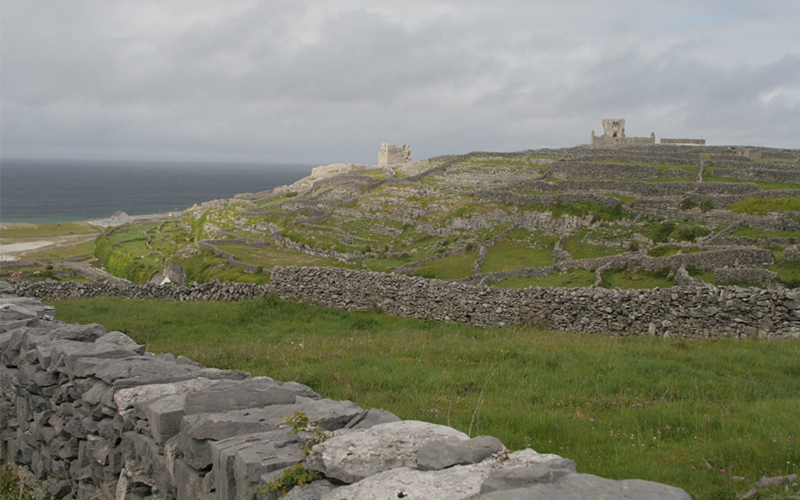 Expansive rural landscape divided by stone walls, with grassy fields and scattered historic stone structures overlooking the coastline. Dark clouds gather above the rocky terrain.