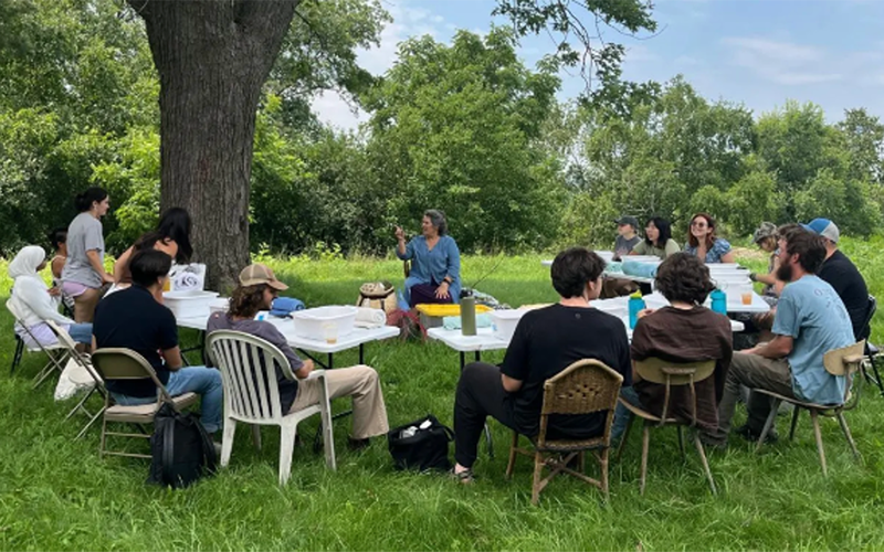 A group of people sits and stands around tables arranged beneath a large tree in a grassy outdoor setting. One participant speaks to the group while others listen during a workshop, discussion, or class session.