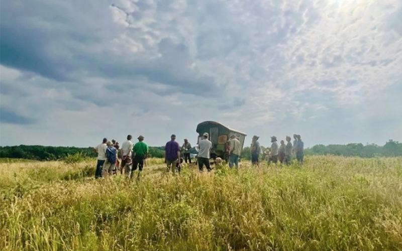 A group of people stands in a grassy field surrounding a covered wagon or mobile structure under a dramatic sky filled with textured clouds.