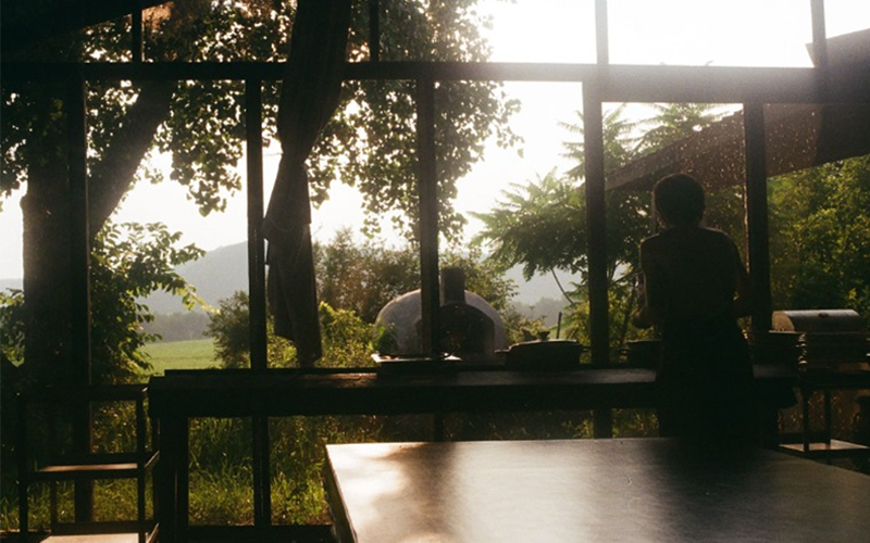 A large window frames a sunlit rural landscape with trees, fields, and distant hills. Interior tables and a standing figure appear in silhouette against the bright outdoor scene.