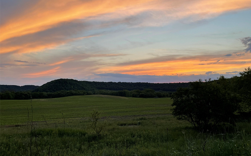 Rolling fields and wooded hills stretch across the horizon beneath a colorful sunset sky streaked with orange, pink, and blue clouds.