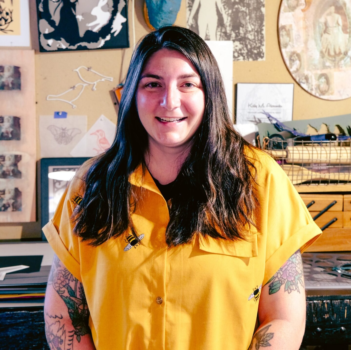A person poses in a creative workspace surrounded by drawings, prints, and artwork pinned to a studio wall.