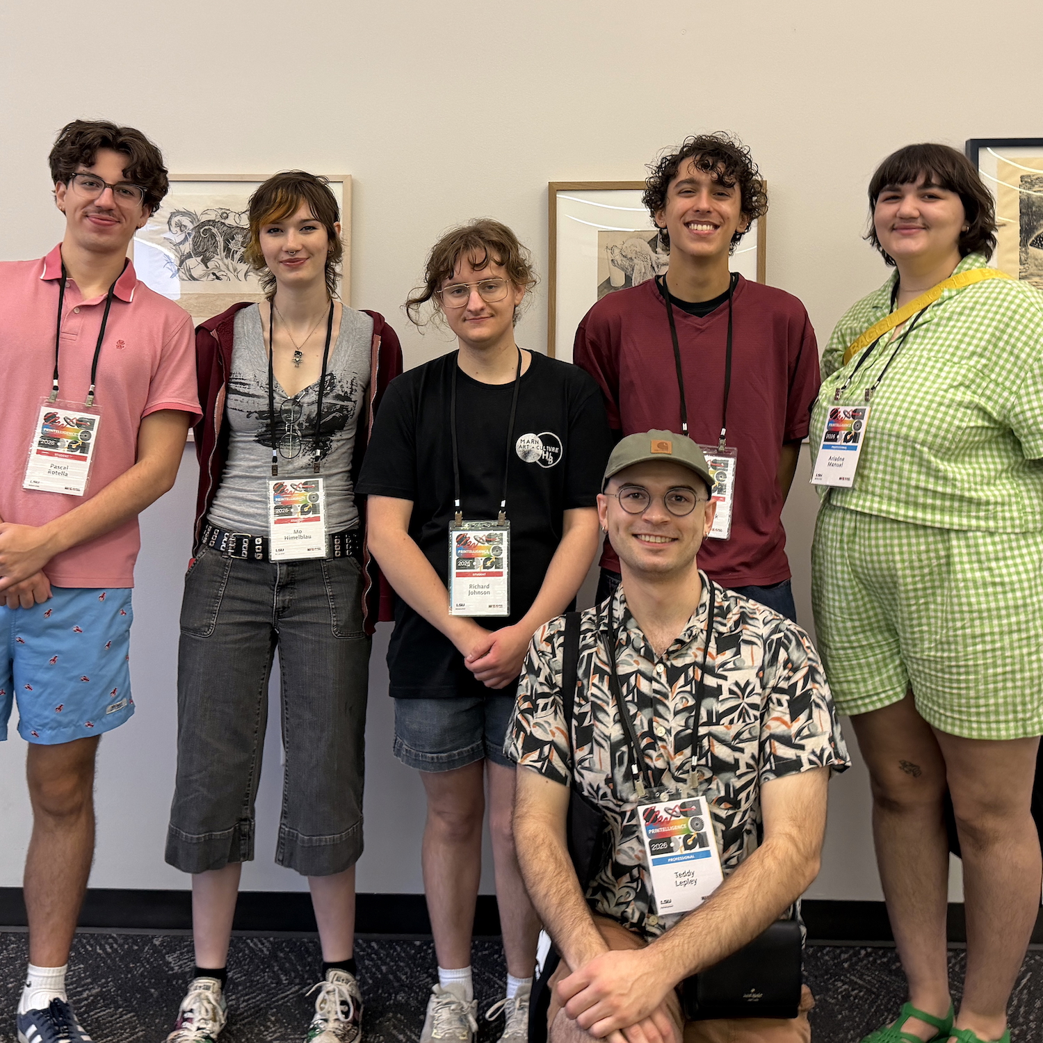 Group of six students wearing event badges posing together indoors in front of framed artwork.